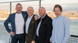Four men standing together outdoors, dressed in casual and formal attire, with a waterfront landscape in the background.