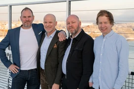 Four men standing together outdoors, dressed in casual and formal attire, with a waterfront landscape in the background.