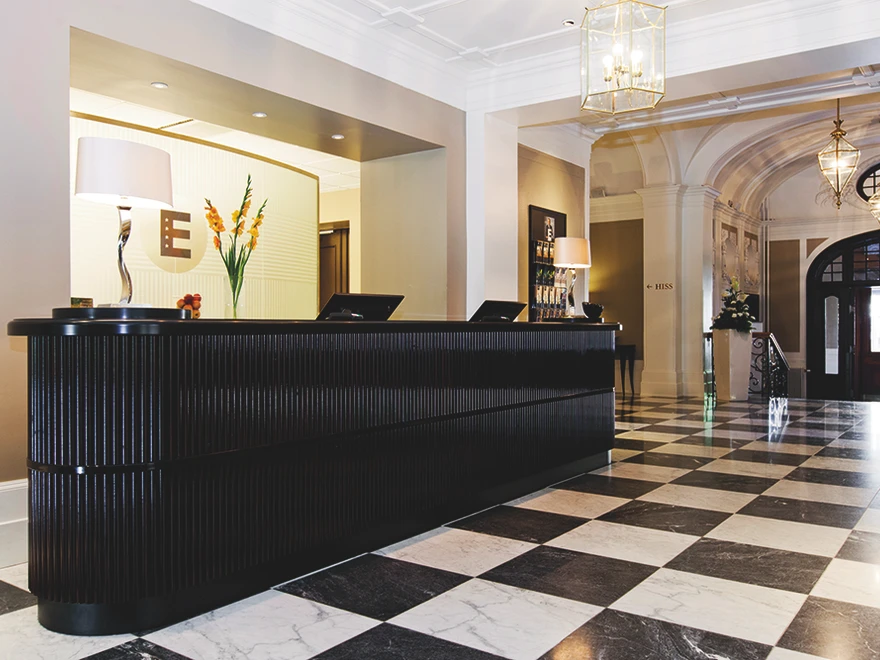 Black reception desk in a well-lit lobby with black-and-white checkered flooring, decorative lamps, and archways.