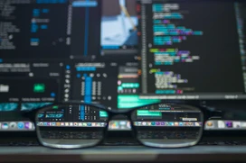 svart datorskärm med rader kod och ett par glasögon som ligger framför 