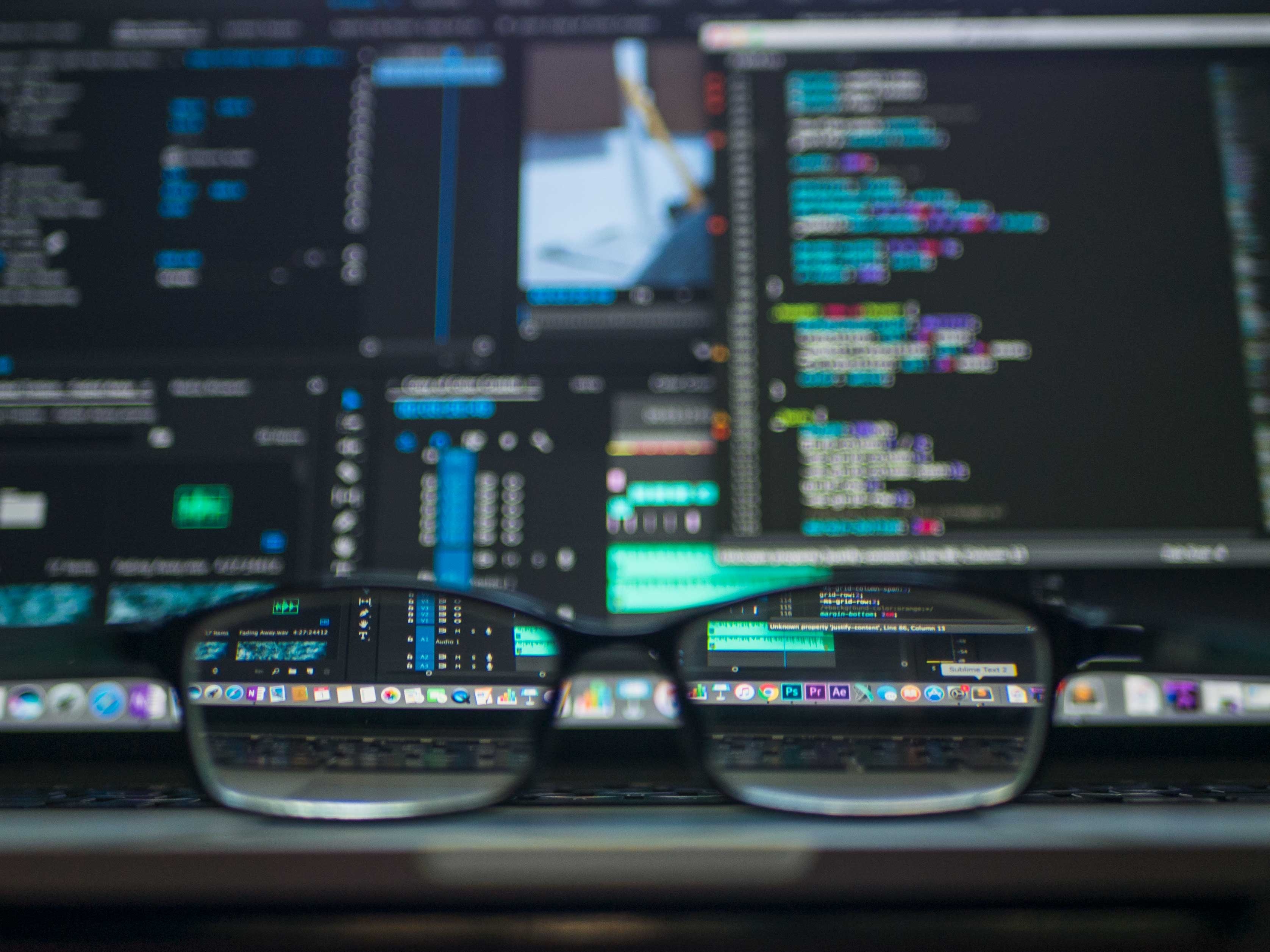svart datorskärm med rader kod och ett par glasögon som ligger framför 