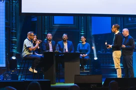 Panel discussion on a conference stage with six blurred speakers, two standing and four seated at a table under blue lighting.