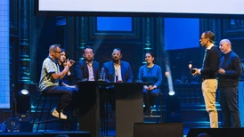 Panel discussion on a conference stage with six blurred speakers, two standing and four seated at a table under blue lighting.