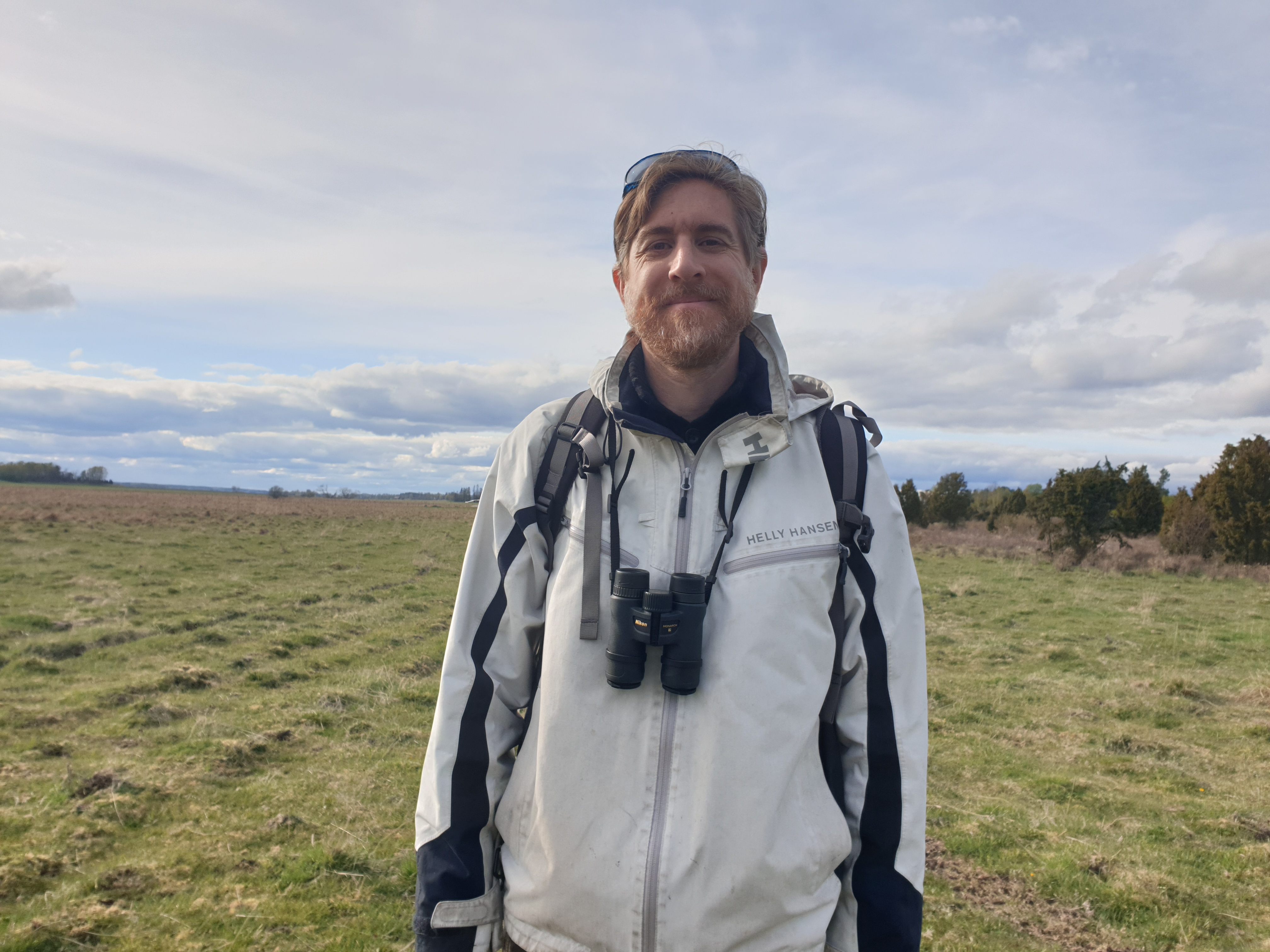 A person standing in an open field wearing a white Helly Hansen jacket, with binoculars around their  neck, under a cloudy sky