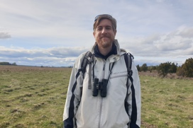 A person standing in an open field wearing a white Helly Hansen jacket, with binoculars around their  neck, under a cloudy sky