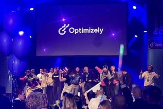 Crowd on stage at an Optimizely event with blue lighting, balloons, and a large presentation screen.
