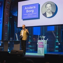 Conference speaker on stage with a large presentation screen showing Anders Borg, blue event lighting, and Optimizely branding.