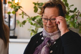 Portrait of a woman in a black polka dot top and purple scarf sitting indoors, with another person partially visible on the left and green plants in the background.