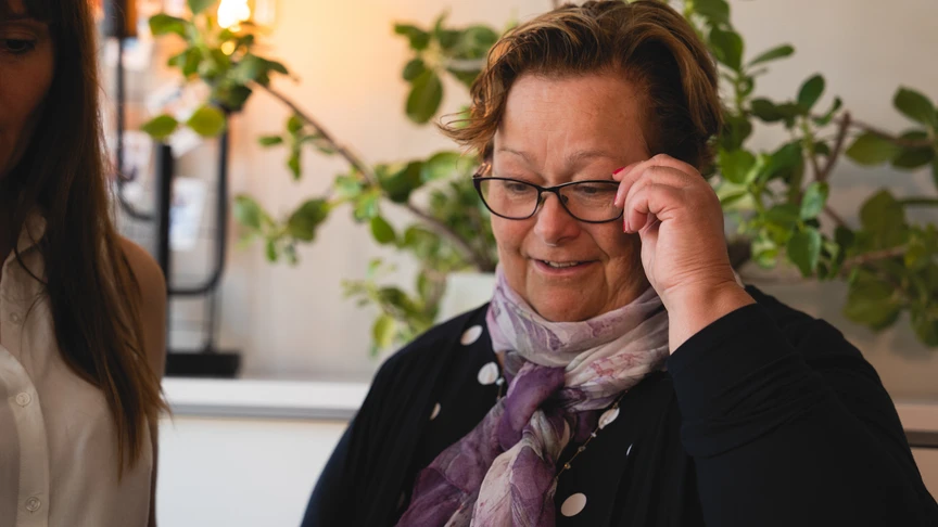 Portrait of a woman in a black polka dot top and purple scarf sitting indoors, with another person partially visible on the left and green plants in the background.