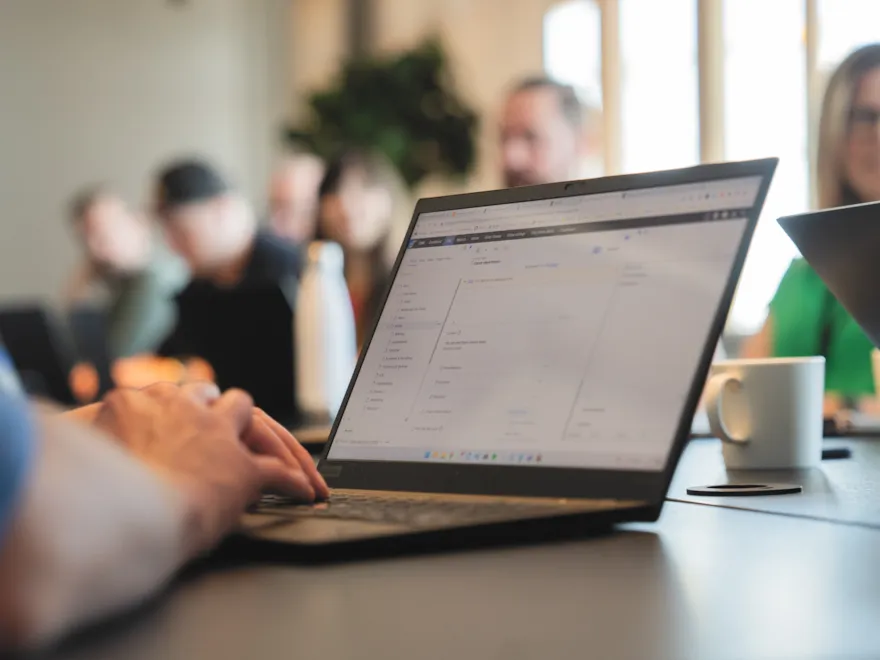 Bärbar datorskärm som visar ett kalkylblad under ett affärsmöte med kollegor i bakgrunden.