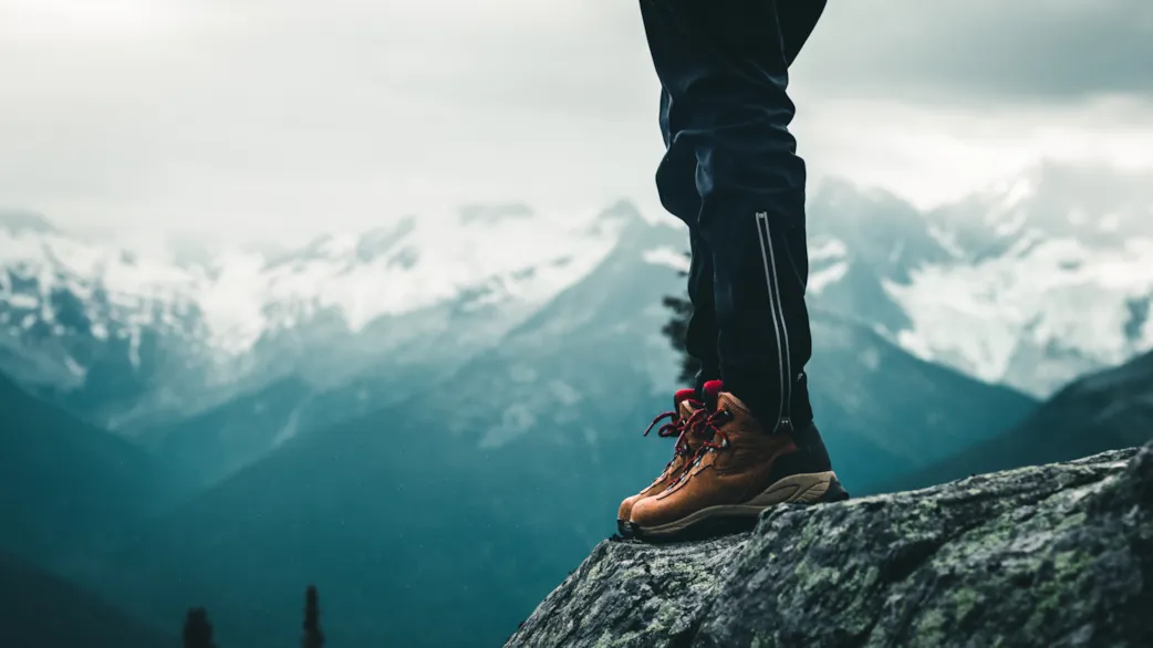 Närbild på en persons fötter och ben – stående högt upp på en klippa med berg i bakgrunden
