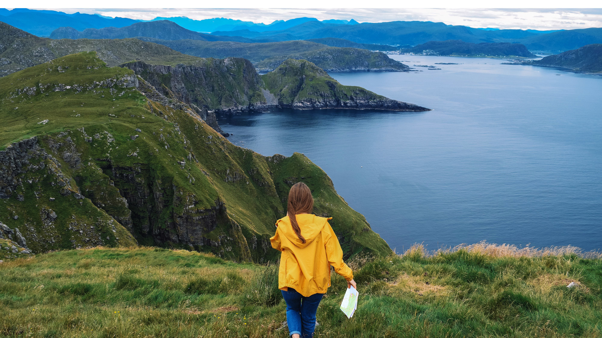 En person i gul jacka håller i en karta med utsikt över fjäll och fjord
